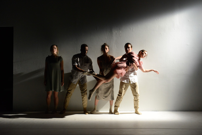 Four dancers stand against a white wall. They hold one women horizontally. She looks like she's floating.